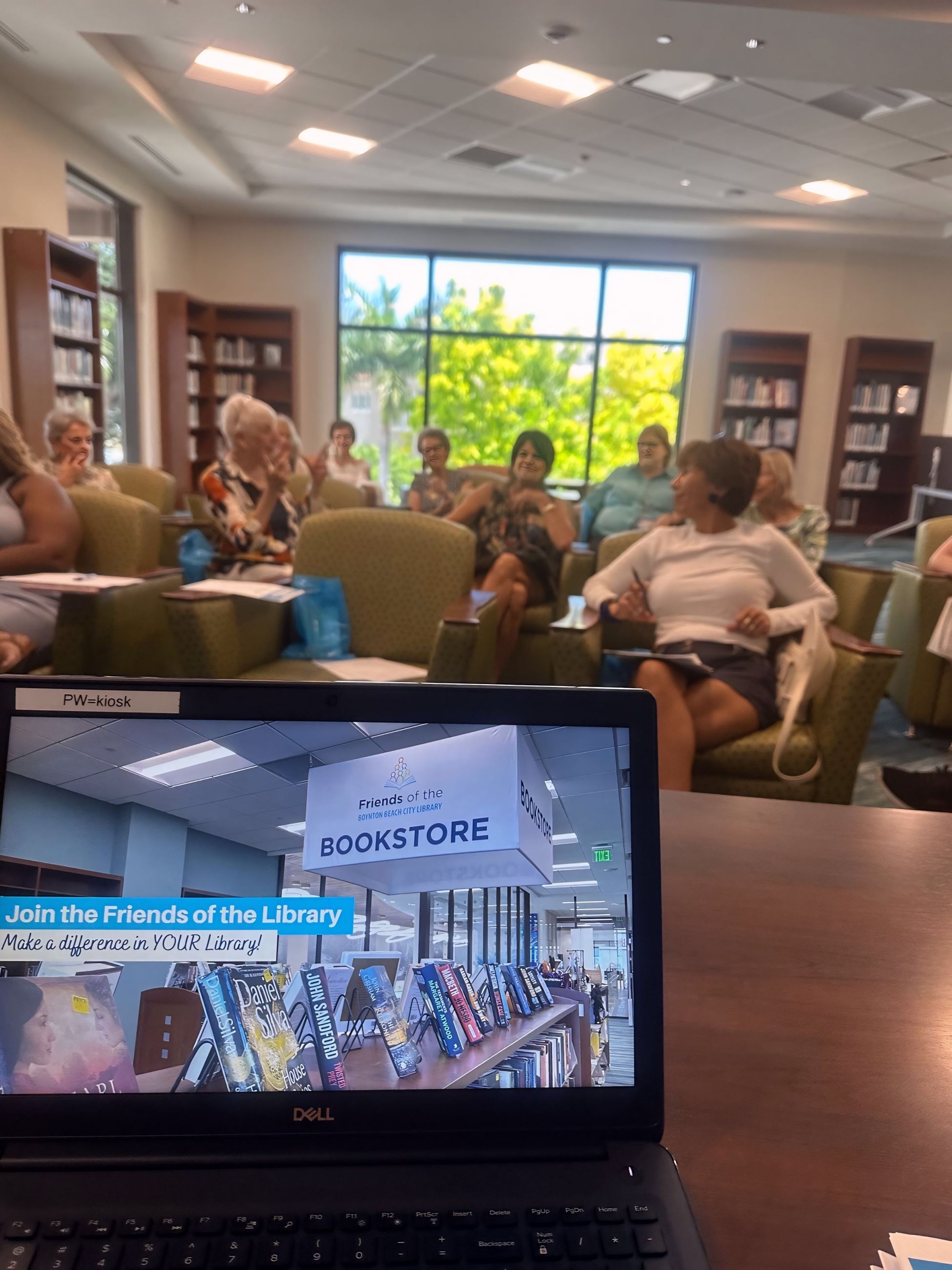 A group of people seated in a library meeting room with bookshelves and large windows showing greene